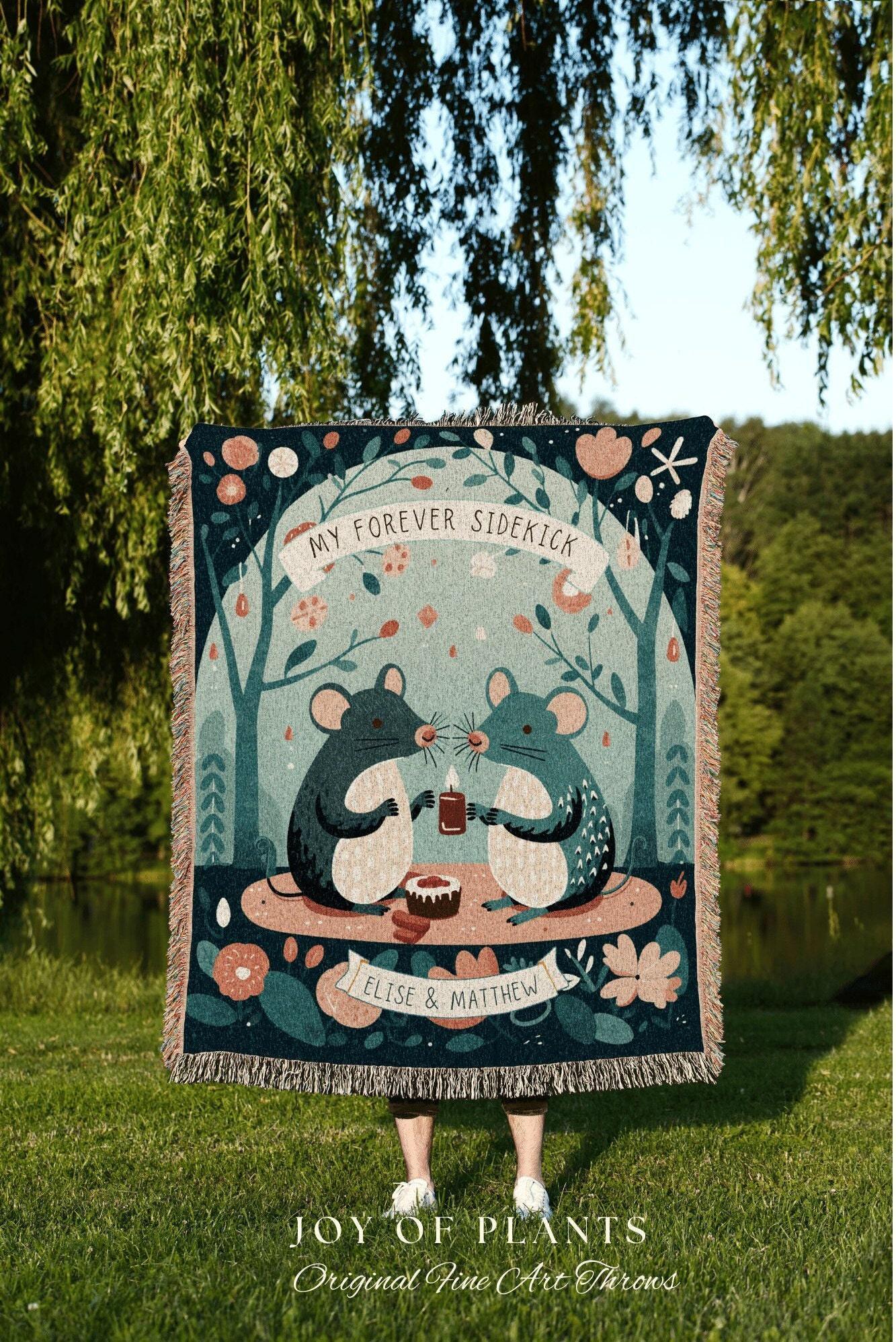 May include: A woven throw blanket with a whimsical illustration of two mice holding a candle, with the text "MY FOREVER SIDEKICK" and "ELISE & MATTHEW". The blanket has a teal background with floral and tree motifs. The bottom of the blanket has a fringe.