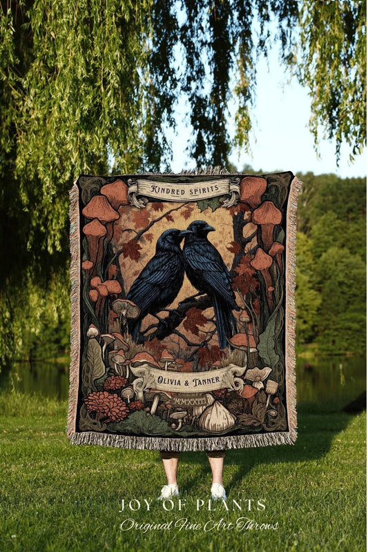May include: A woven throw blanket featuring a nature-themed design with two black ravens perched on a branch. The blanket has a fringe border and includes text that reads "Kindred Spirits" and "Olivia & Tanner". The bottom of the blanket reads "JOY OF PLANTS Original Fine Art Throws".