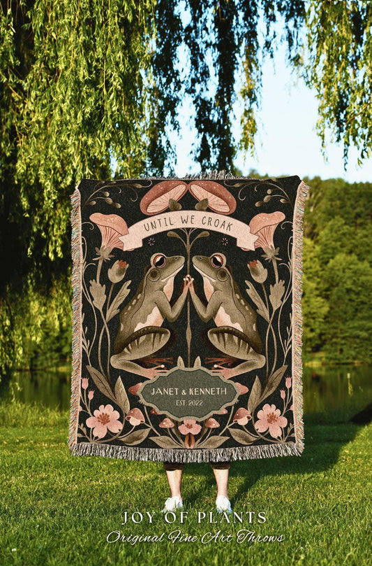 May include: A black throw blanket featuring two frogs facing each other, with the text "UNTIL WE CROAK" above them. The blanket has a floral and mushroom design, with the names "JANET & KENNETH" and the year "EST. 2022" below the frogs. The bottom of the blanket has a fringe.