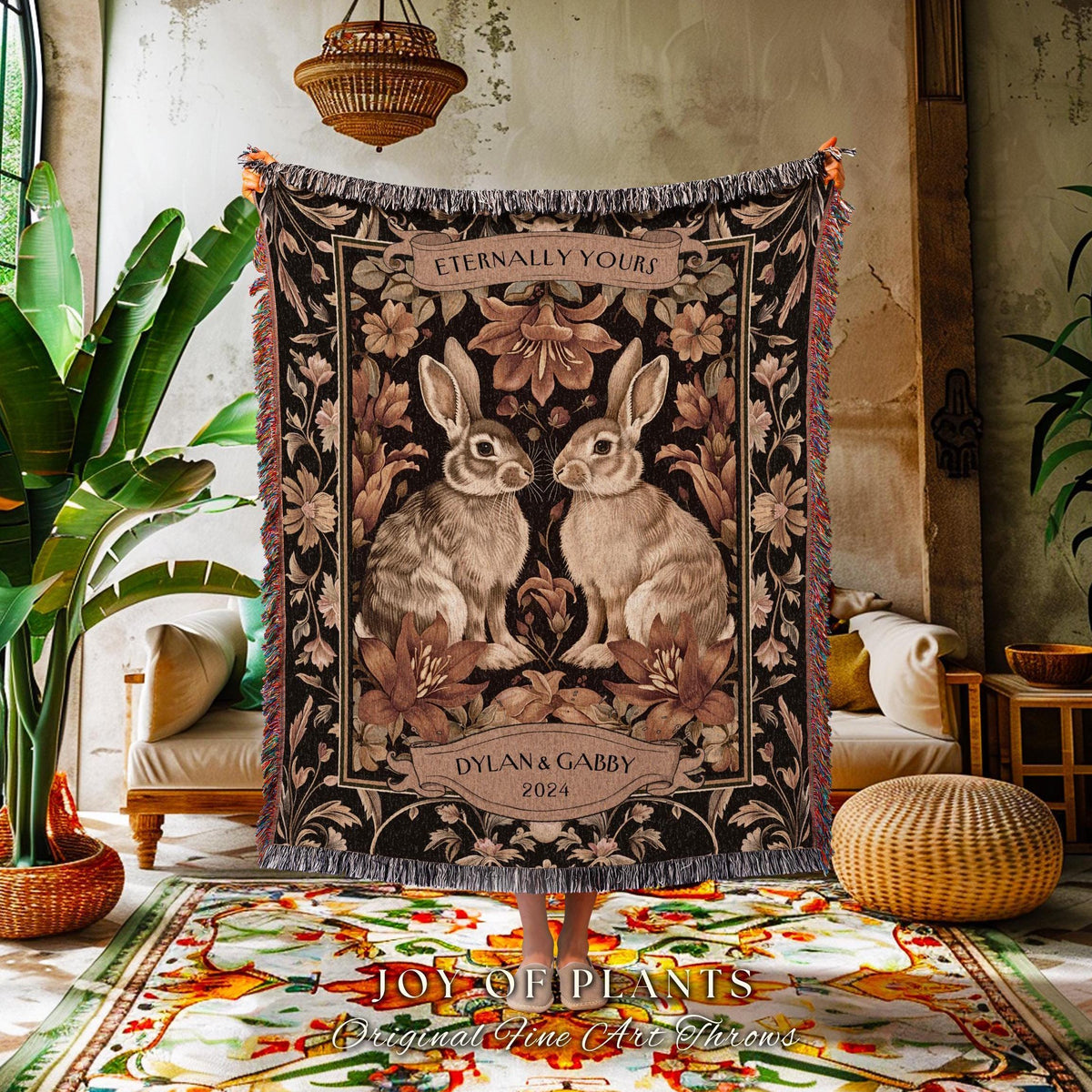 May include: A woven throw blanket featuring two rabbits facing each other, surrounded by floral designs in shades of brown and beige. The text "Eternally Yours" is at the top, with "Dylan & Gabby 2024" below. The blanket has a fringed edge.
