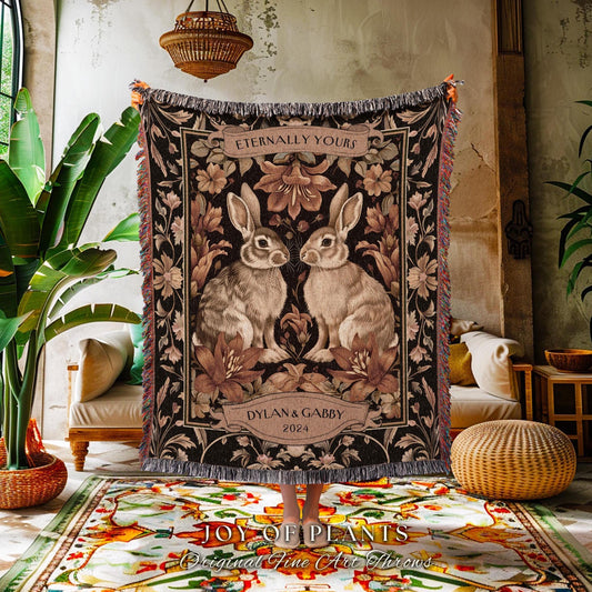 May include: A woven throw blanket featuring two rabbits facing each other, surrounded by floral designs in shades of brown and beige. The text "Eternally Yours" is at the top, with "Dylan & Gabby 2024" below. The blanket has a fringed edge.