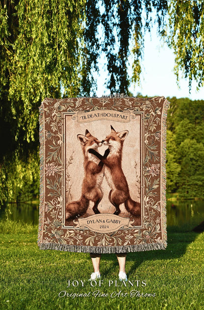 May include: A brown throw blanket featuring two foxes kissing, framed by floral and scroll designs. The text "TIL DEATH DO US PART" and "DYLAN & GABBY 2024" are displayed. The blanket has a fringed edge and is held outdoors.