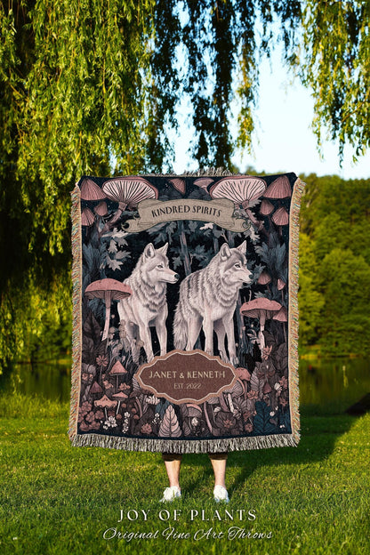 May include: A woven throw blanket featuring a forest scene with two wolves, mushrooms, and the text "KINDRED SPIRITS". The blanket has a dark background with a beige fringe and the text "JANET & KENNETH EST. 2022".