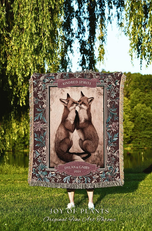 May include: A decorative throw blanket featuring a sepia-toned image of two foxes touching noses. The blanket has a dark brown border with floral accents and the words "Kindred Spirits" and "Dylan & Gabby 2024". The bottom of the blanket reads "JOY OF PLANTS Original Fine Art Throws".