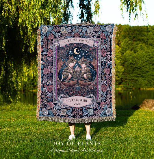 May include: A tapestry throw blanket with a detailed illustration of two frogs wearing crowns, beneath a crescent moon. The blanket features the text "UNTIL WE CROAK" and "DYLAN & GABBY 2025". The blanket has a floral border and fringe.