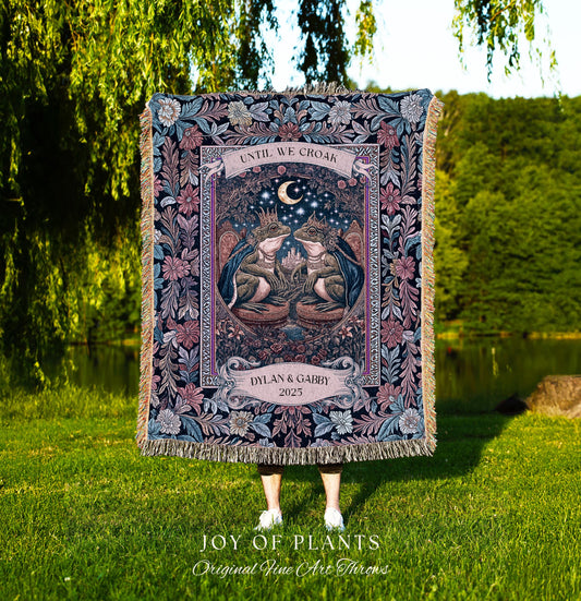May include: A tapestry throw blanket with a detailed illustration of two frogs wearing crowns, beneath a crescent moon. The blanket features the text "UNTIL WE CROAK" and "DYLAN & GABBY 2025". The blanket has a floral border and fringe.