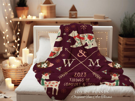 May include: A burgundy throw blanket with a festive design featuring a bear, deer, and owl. The blanket has the text "2023 Tidings of Comfort & Joy" and the initials "W & M". The blanket is on a white bed.