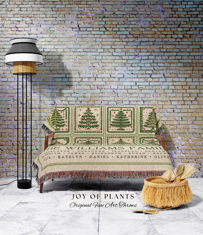 May include: A beige throw blanket with green Christmas tree designs and the text "THE WILLIAMS FAMILY". The blanket is draped over a wooden sofa. A floor lamp and a woven basket are also in the image.