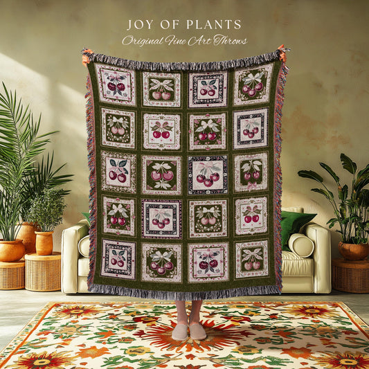 May include: A green throw blanket with a cherry pattern. The blanket has a fringe and is held up by a person. The text "JOY OF PLANTS Original Fine Art Throws" is at the top.