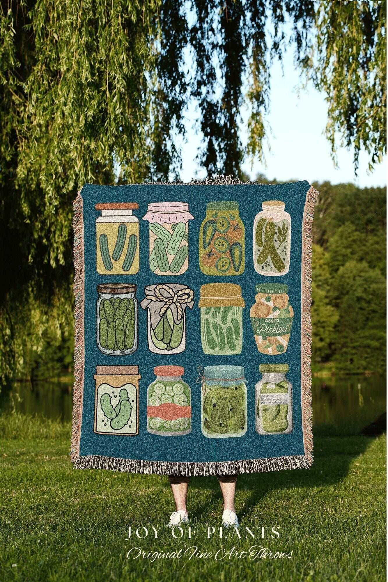 May include: A teal throw blanket featuring illustrations of various pickle jars. The jars are filled with pickles and other ingredients. The blanket has a fringed edge and the text "JOY OF PLANTS Original Fine Art Throws" at the bottom.