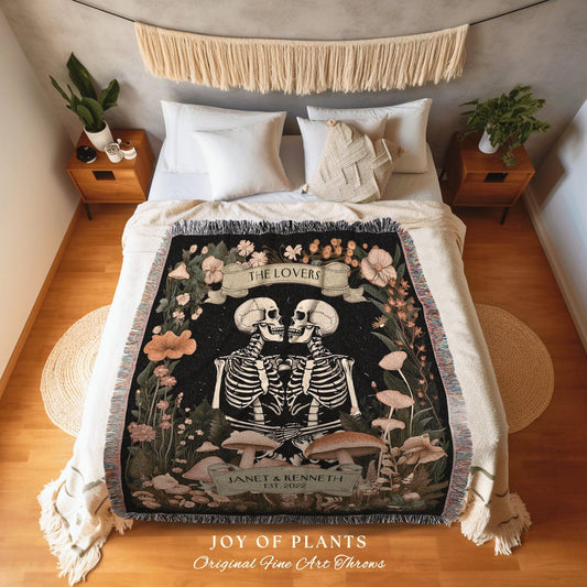 May include: A black throw blanket featuring a skeleton couple design with the text "THE LOVERS" and "JANET & KENNETH EST. 2022". The blanket has a floral and mushroom motif with fringe edges. The words "JOY OF PLANTS" and "Original Fine Art Throws" are also visible.