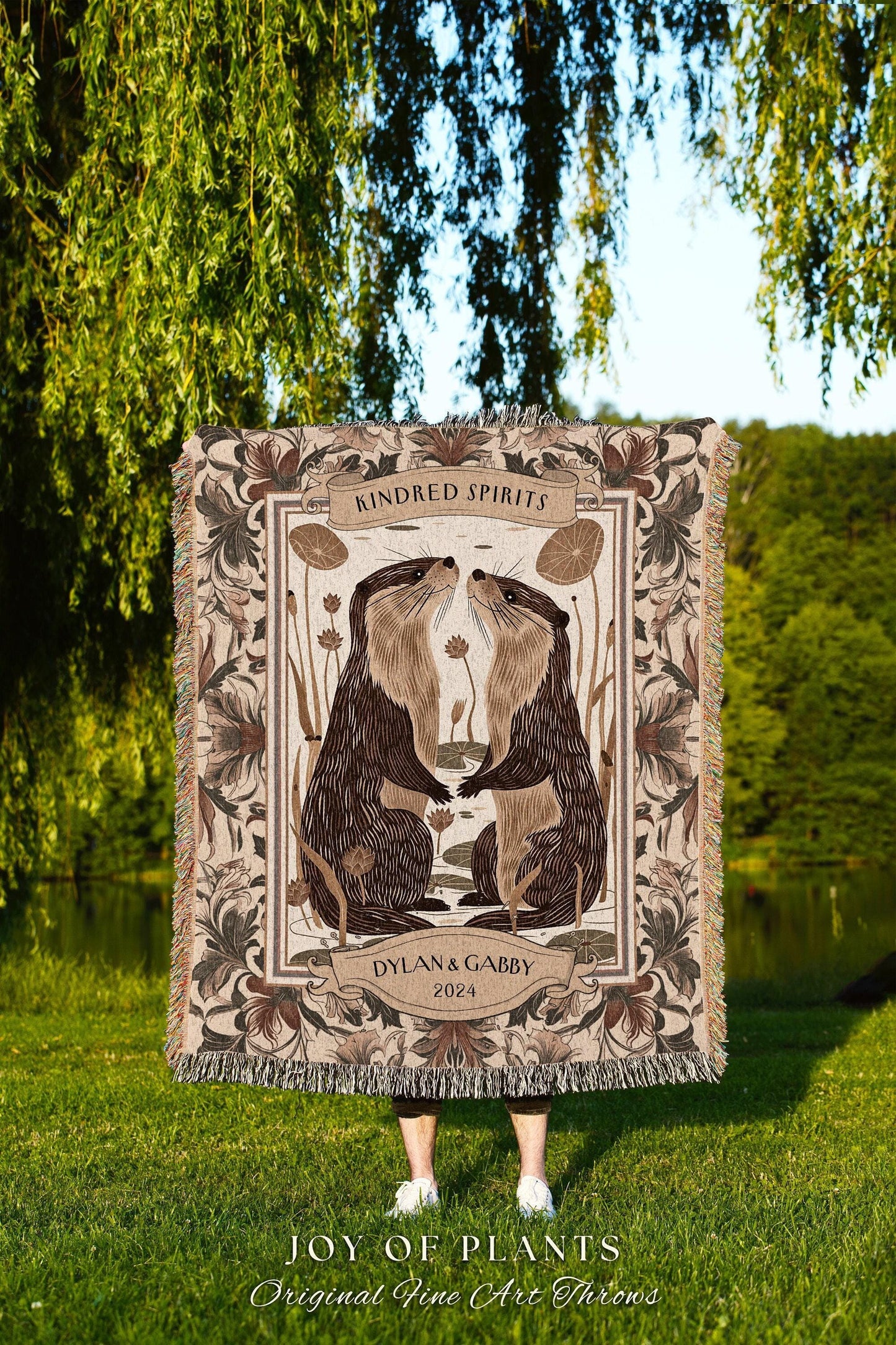 May include: A woven throw blanket featuring an illustration of two otters touching noses, with the words "Kindred Spirits" and "Dylan & Gabby 2024". The blanket has a floral border and fringe.