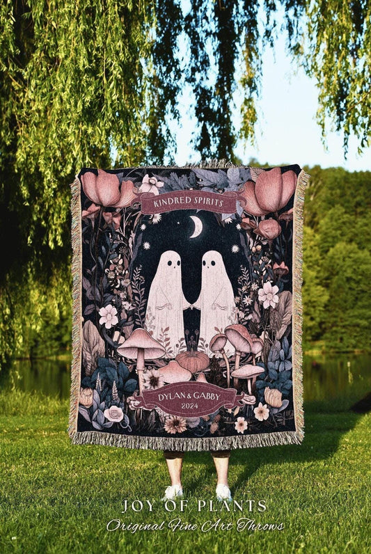 May include: A woven throw blanket featuring a whimsical design with two ghost figures holding hands, surrounded by mushrooms and flowers. The words "Kindred Spirits" and "Dylan & Gabby 2024" are displayed on the blanket. The blanket has a fringed edge.