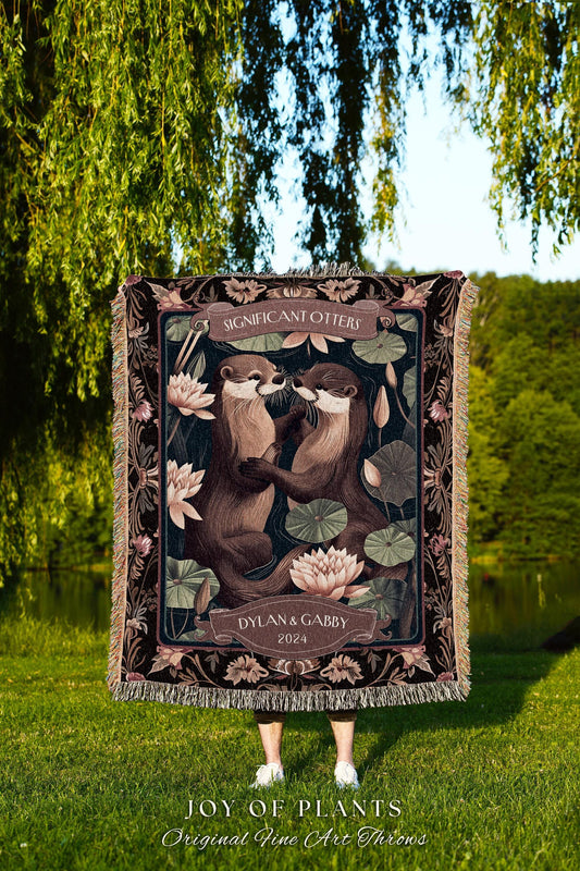 May include: A woven throw blanket featuring two otters embracing, surrounded by lotus flowers and foliage. The blanket has a dark background with the text "SIGNIFICANT OTTERS" and "DYLAN & GABBY 2024". The bottom of the blanket reads "JOY OF PLANTS Original Fine Art Throws".
