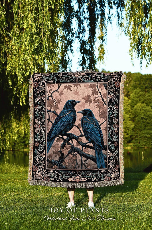 May include: A woven throw blanket featuring two blue-black ravens perched on a tree branch. The blanket has a decorative border with floral and leaf patterns. The background is a soft pink with tree branches and leaves. The text "JOY OF PLANTS Original Fine Art Throws" is at the bottom.