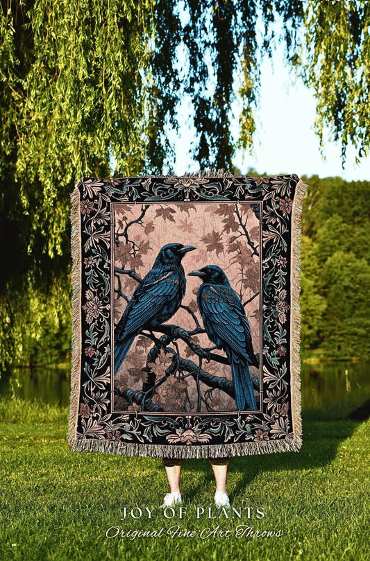 May include: A woven throw blanket featuring two blue-black ravens perched on a tree branch. The blanket has a decorative border with floral and leaf patterns. The background is a soft pink with tree branches and leaves. The text "JOY OF PLANTS Original Fine Art Throws" is at the bottom.