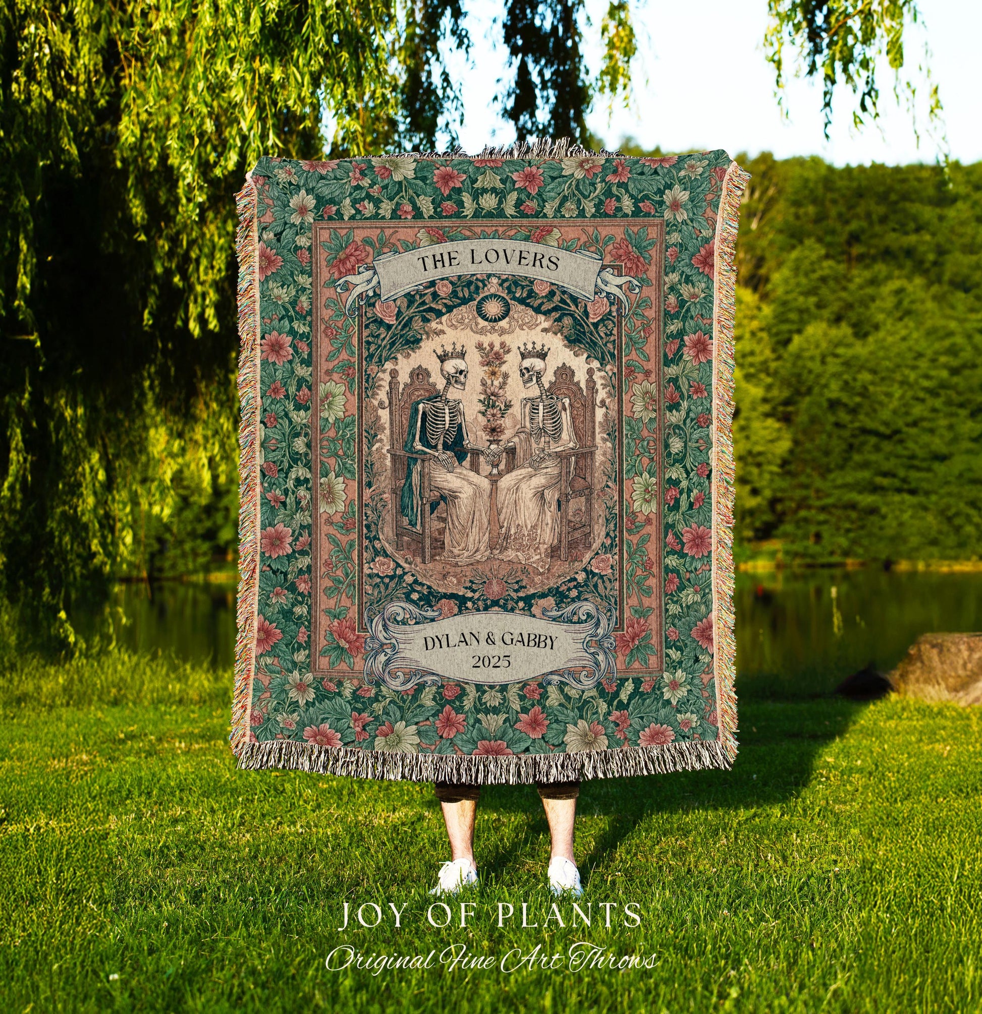 May include: A tapestry throw blanket with a floral border and a central image of two skeletons seated on thrones. The banner reads "THE LOVERS" above the image, and "DYLAN & GABBY 2025" below. The blanket is fringed.