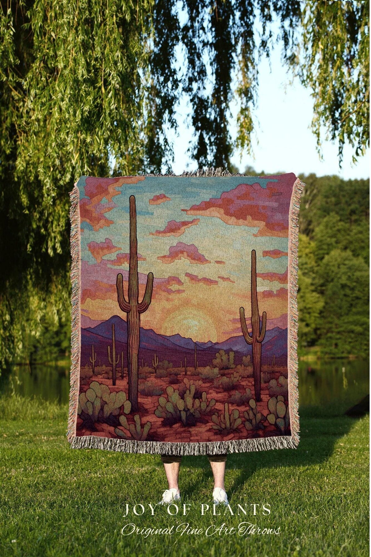 May include: A woven throw blanket featuring a desert landscape with cacti, mountains, and a sunset. The blanket has a fringed edge and is held up in front of a person. The text "JOY OF PLANTS Original Fine Art Throws" is visible.
