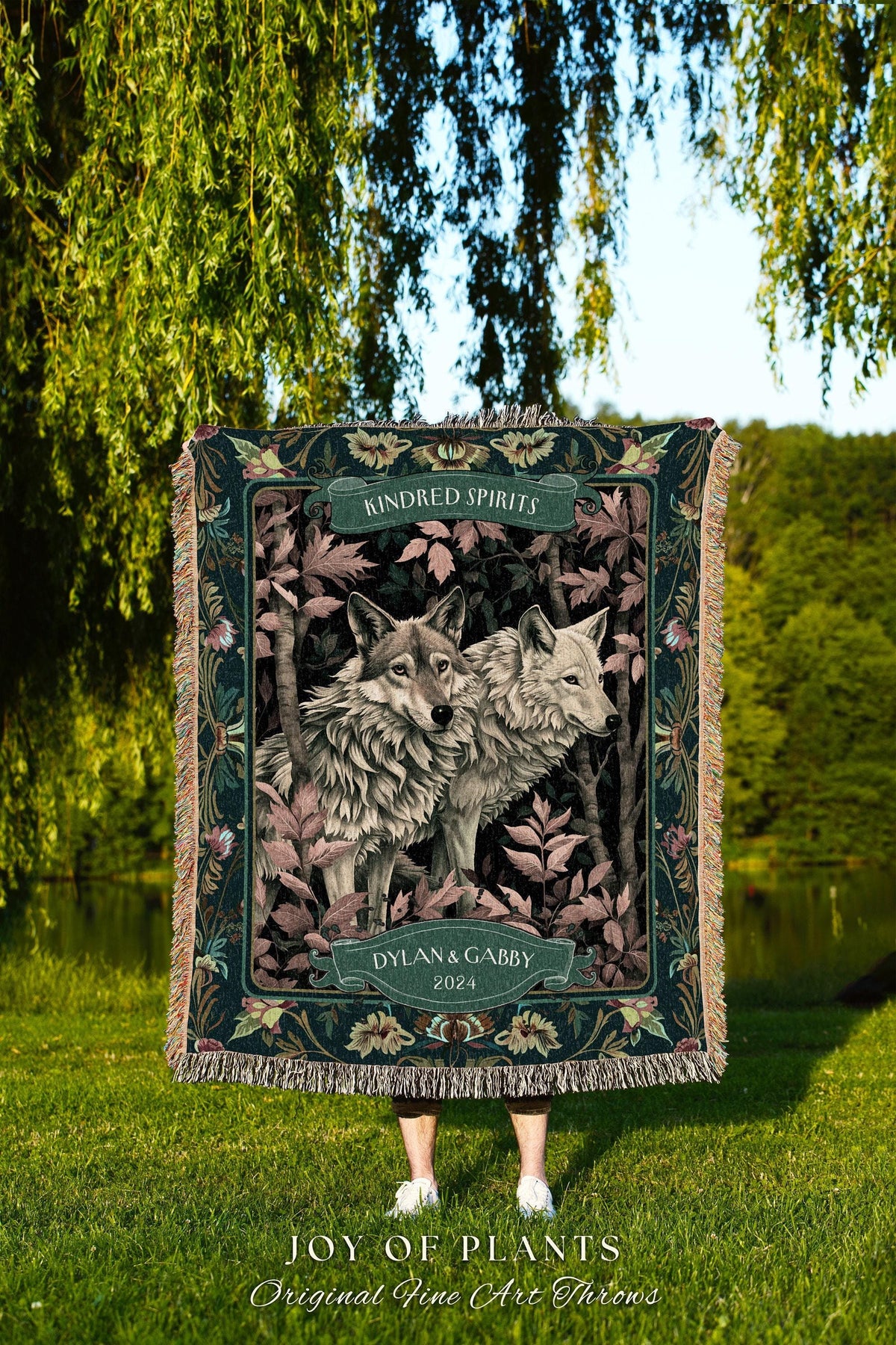 May include: A decorative throw blanket featuring two wolves in a forest setting. The blanket has a dark green border with floral accents and the words "Kindred Spirits" and "Dylan & Gabby 2024". The bottom of the blanket reads "JOY OF PLANTS Original Fine Art Throws".