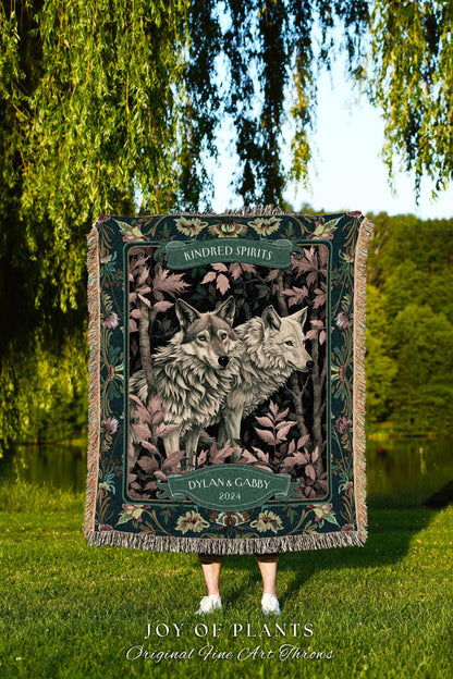May include: A decorative throw blanket featuring two wolves in a forest setting. The blanket has a dark green border with floral accents and the words "Kindred Spirits" and "Dylan & Gabby 2024". The bottom of the blanket reads "JOY OF PLANTS Original Fine Art Throws".