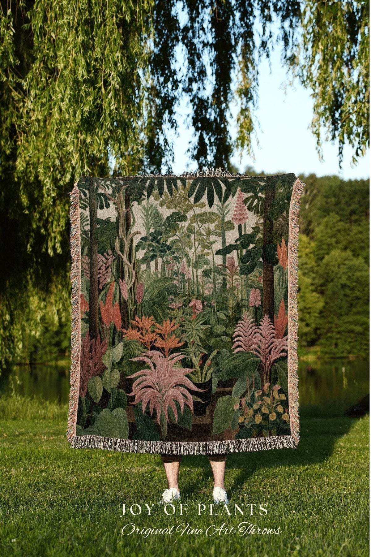 May include: A woven throw blanket featuring a botanical illustration of various plants in shades of green, pink, and orange. The blanket has a fringed edge and the text "JOY OF PLANTS Original Fine Art Throws" at the bottom.