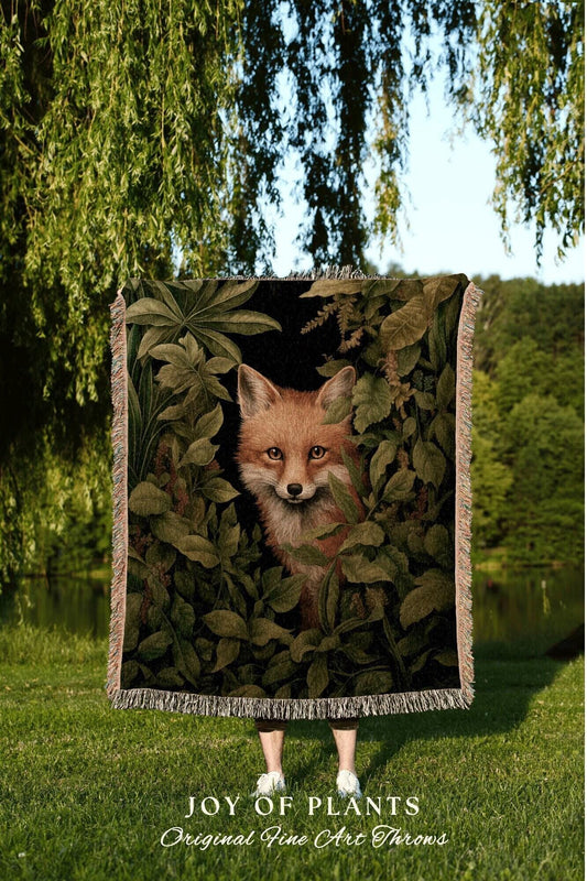 May include: A woven throw blanket featuring a fox peering out from lush green foliage. The artwork has a dark background with a fox in the center. The blanket has a fringed edge and the text "JOY OF PLANTS Original Fine Art Throws" at the bottom.