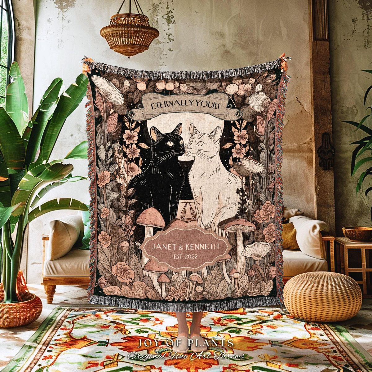 May include: A decorative throw blanket featuring a black and white cat illustration with the text "Eternally Yours" and "Janet & Kenneth Est. 2022". The blanket has a floral and mushroom design in shades of brown, beige, and black.