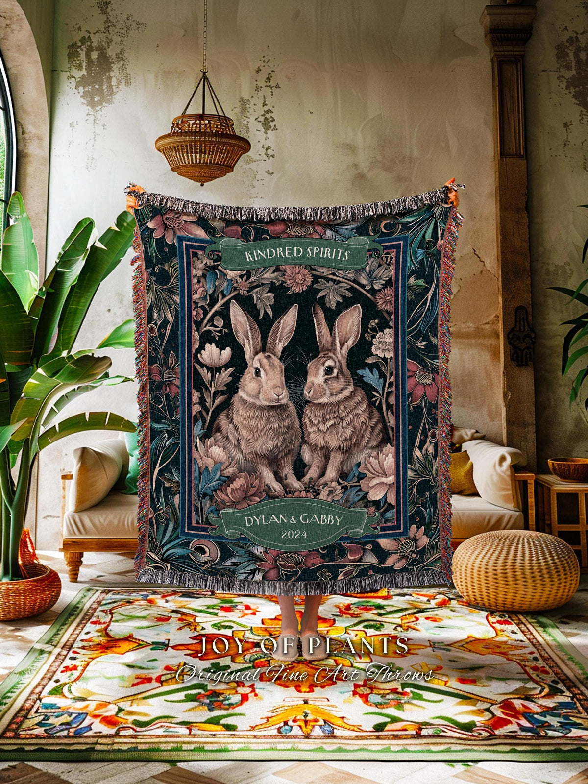 May include: A dark blue throw blanket featuring two rabbits surrounded by flowers. The blanket has a decorative border and fringe. Text on the blanket reads "Kindred Spirits" and "Dylan & Gabby 2024". The words "JOY OF PLANTS" and "Original Fine Art Throws" are at the bottom.