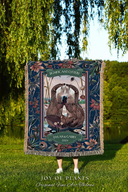 May include: A woven throw blanket featuring a detailed illustration of two otters touching noses, framed by floral and botanical patterns in shades of blue, green, and brown. The text "SIGNIFICANT OTTERS" and "DYLAN & GABBY 2024" are displayed on the blanket.