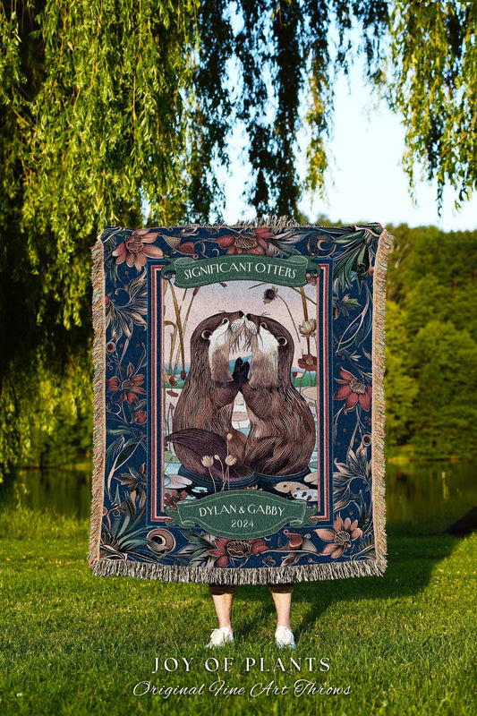 May include: A woven throw blanket featuring a detailed illustration of two otters touching noses, framed by floral and botanical patterns in shades of blue, green, and brown. The text "SIGNIFICANT OTTERS" and "DYLAN & GABBY 2024" are displayed on the blanket.