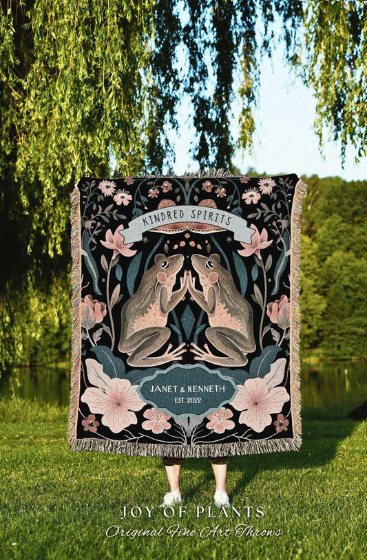 May include: A black throw blanket with a floral and frog design. The blanket features two frogs touching hands, surrounded by flowers and foliage. Text includes "Kindred Spirits" and "Janet & Kenneth EST. 2022". The blanket has a fringed edge.