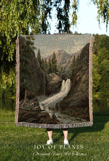 May include: A woven throw blanket featuring a detailed landscape of a waterfall cascading down a rocky cliff, surrounded by trees. The blanket has a fringed edge and the text "JOY OF PLANTS Original Fine Art Throws" at the bottom.