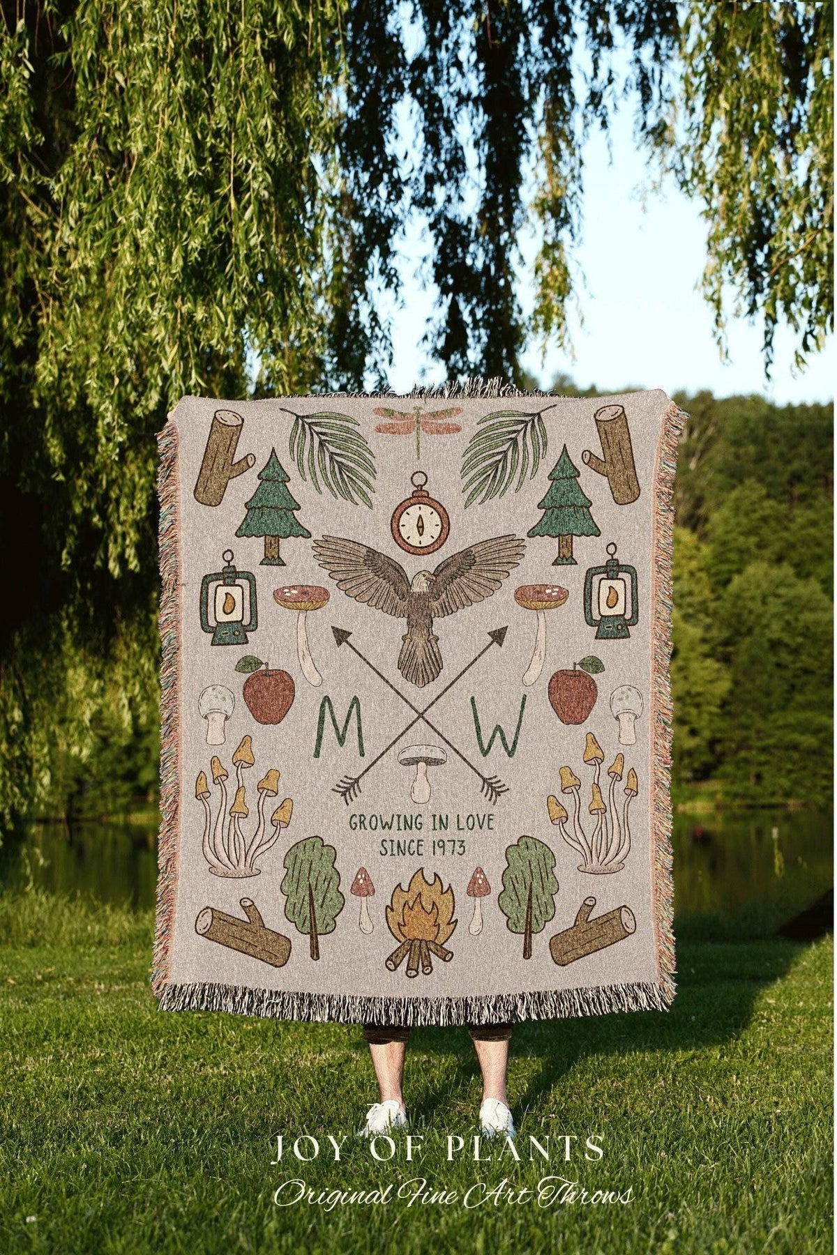 May include: A beige throw blanket with a nature-themed design. The blanket features illustrations of trees, mushrooms, a compass, an eagle, and the text "Growing in Love Since 1973". The blanket has a fringed edge.