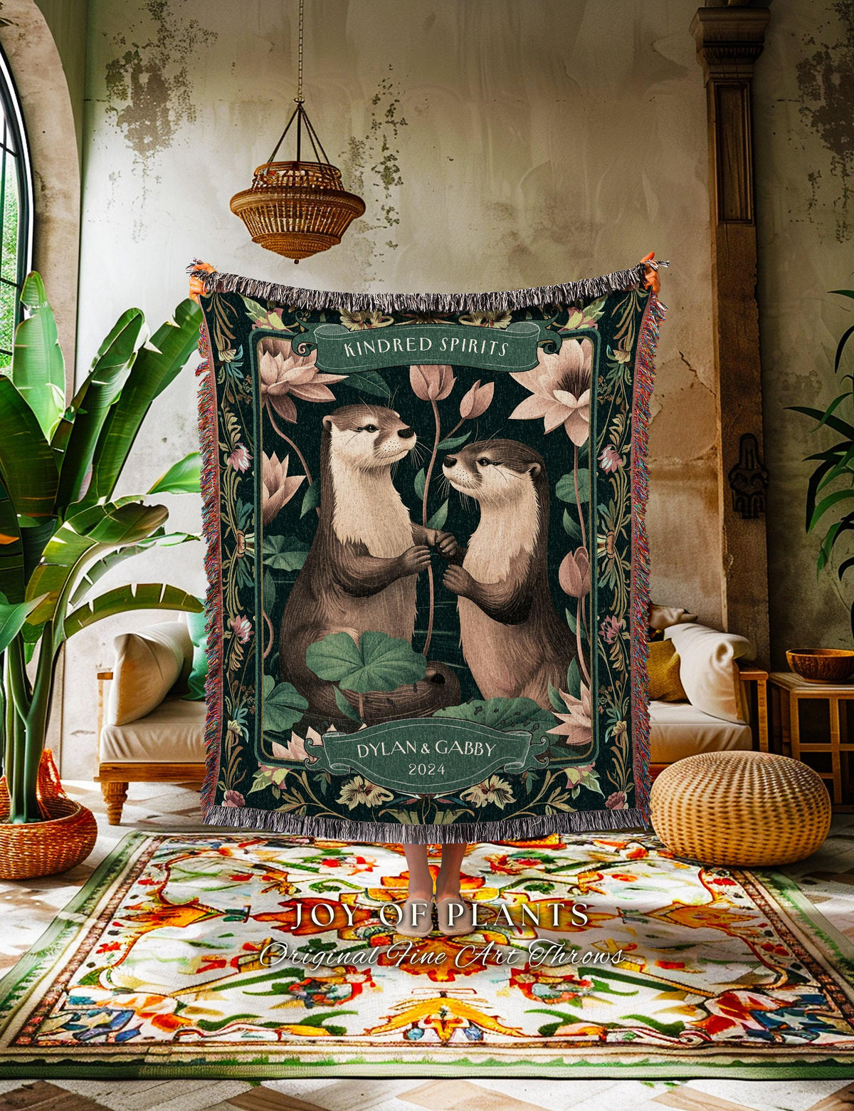 May include: A dark green throw blanket featuring two otters holding hands, surrounded by lotus flowers. The blanket has the text "Kindred Spirits" and "Dylan & Gabby 2024". The blanket has a fringed edge and is displayed in a room with plants and a patterned rug.