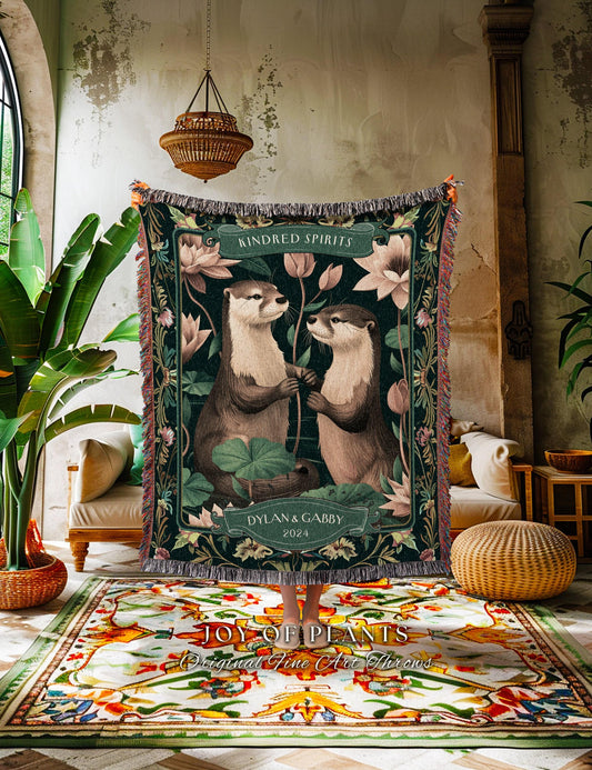 May include: A dark green throw blanket featuring two otters holding hands, surrounded by lotus flowers. The blanket has the text "Kindred Spirits" and "Dylan & Gabby 2024". The blanket has a fringed edge and is displayed in a room with plants and a patterned rug.
