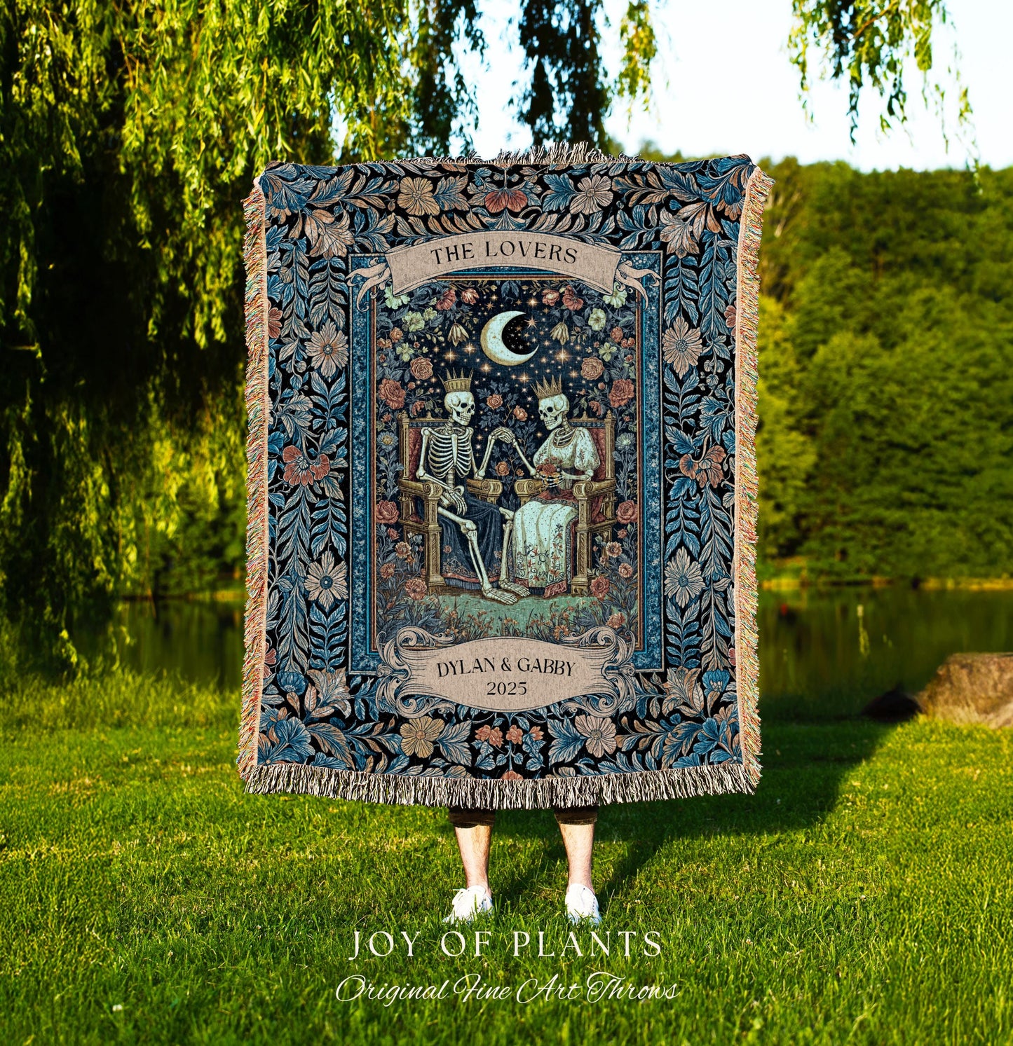 May include: A dark blue tapestry throw blanket with a floral and skeleton design. The blanket features the text "THE LOVERS" and "DYLAN & GABBY 2025". The throw has a fringed edge and is displayed outdoors.