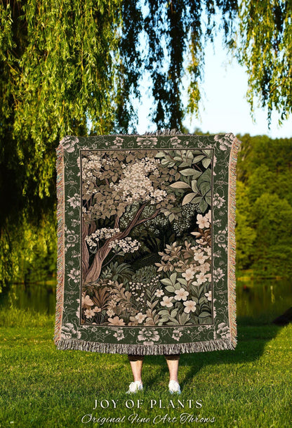 May include: A woven throw blanket featuring a detailed forest scene with trees, flowers, and foliage in shades of green, brown, and white. The blanket has a dark green border with floral patterns and fringe. The text "JOY OF PLANTS" is visible.