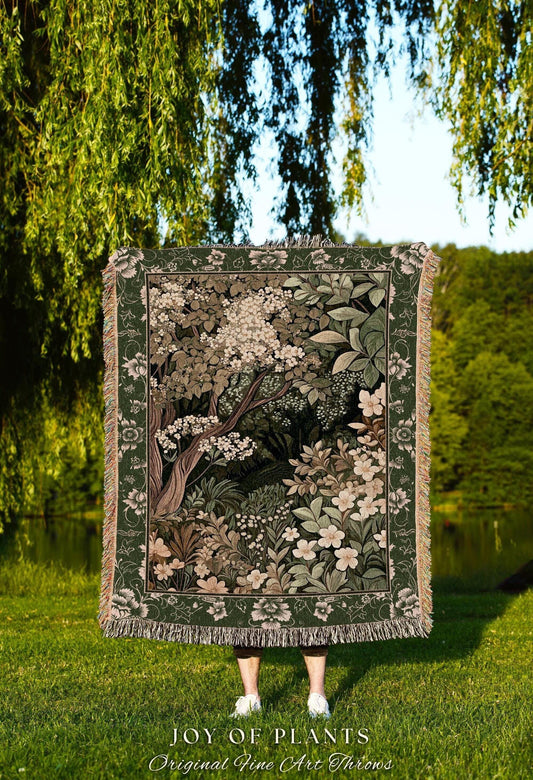 May include: A woven throw blanket featuring a detailed forest scene with trees, flowers, and foliage in shades of green, brown, and white. The blanket has a dark green border with floral patterns and fringe. The text "JOY OF PLANTS" is visible.