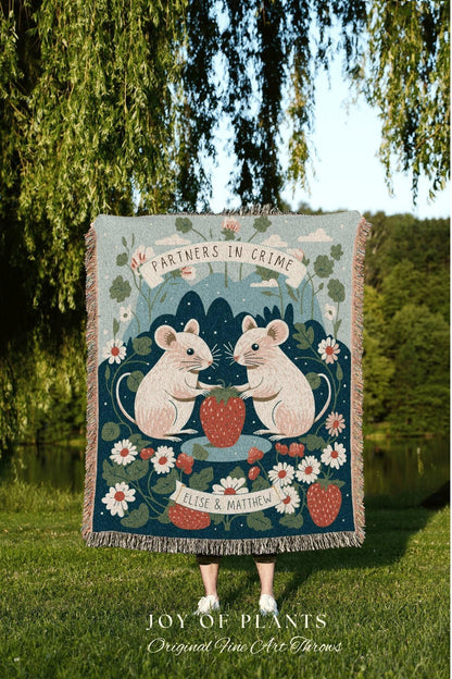 May include: A woven throw blanket featuring a whimsical design with two mice holding a strawberry, surrounded by flowers. The text "PARTNERS IN CRIME" and names are displayed on the blanket. The blanket has a fringed edge.